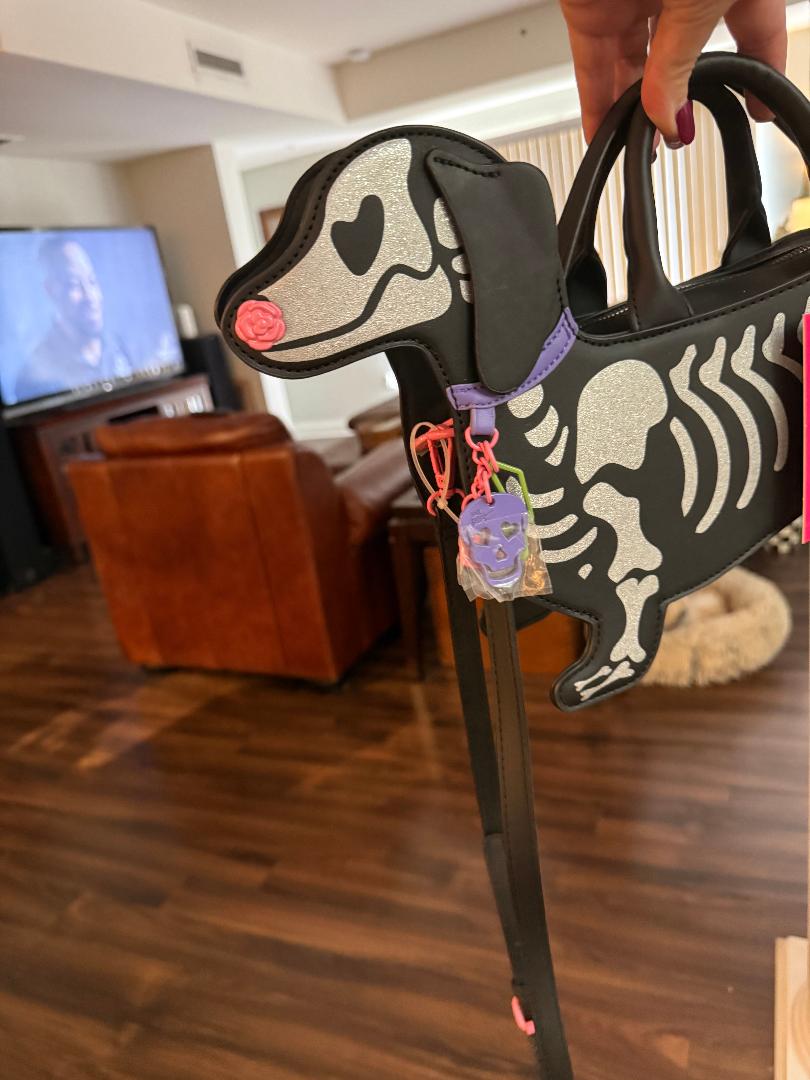 A black and silver glitter dachshund-shaped handbag with a skeleton design, held by a hand in a living room setting.