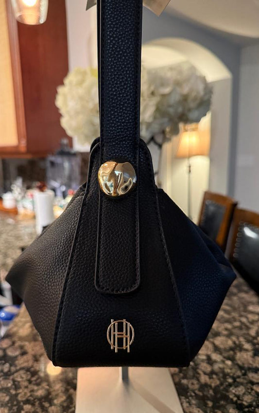 A black pebbled leather handbag with a structured geometric shape, a single shoulder strap, and gold-tone hardware, displayed on a stand in a kitchen setting.