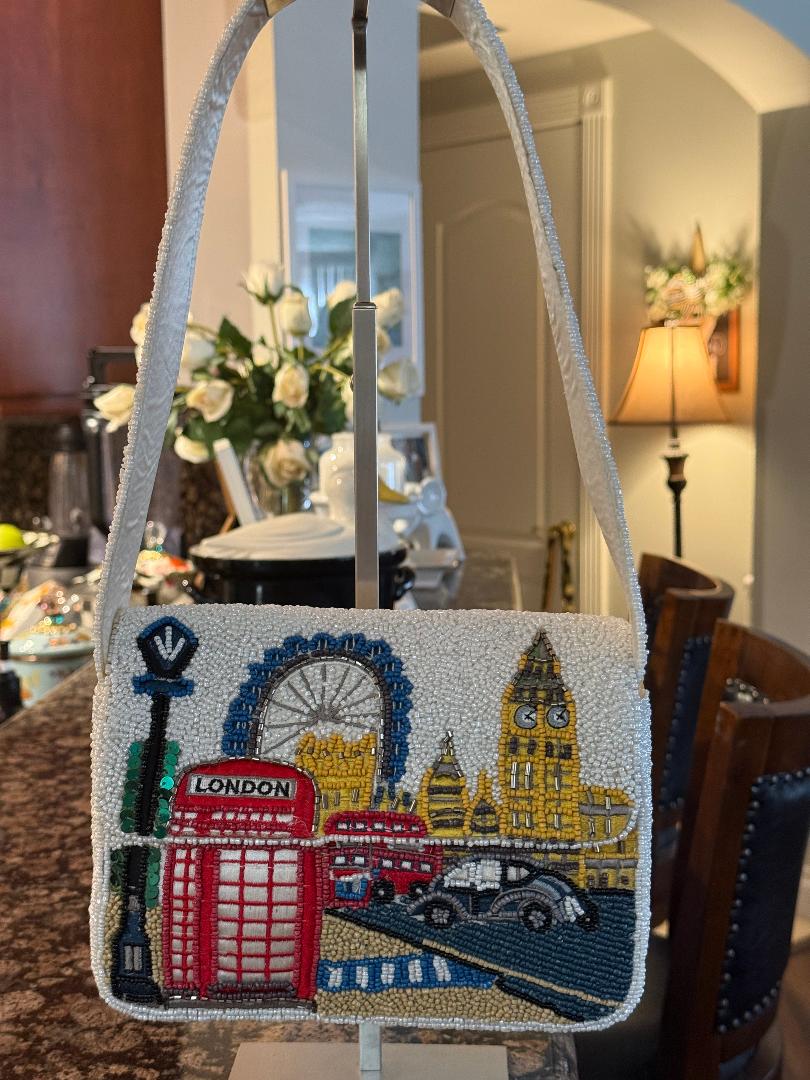 A beaded London-themed shoulder bag with white faux fur trim, featuring embroidered red double-decker buses, a red telephone booth, Big Ben, the London Eye, and a black car on a street scene. The bag is displayed on a stand in a home interior setting.