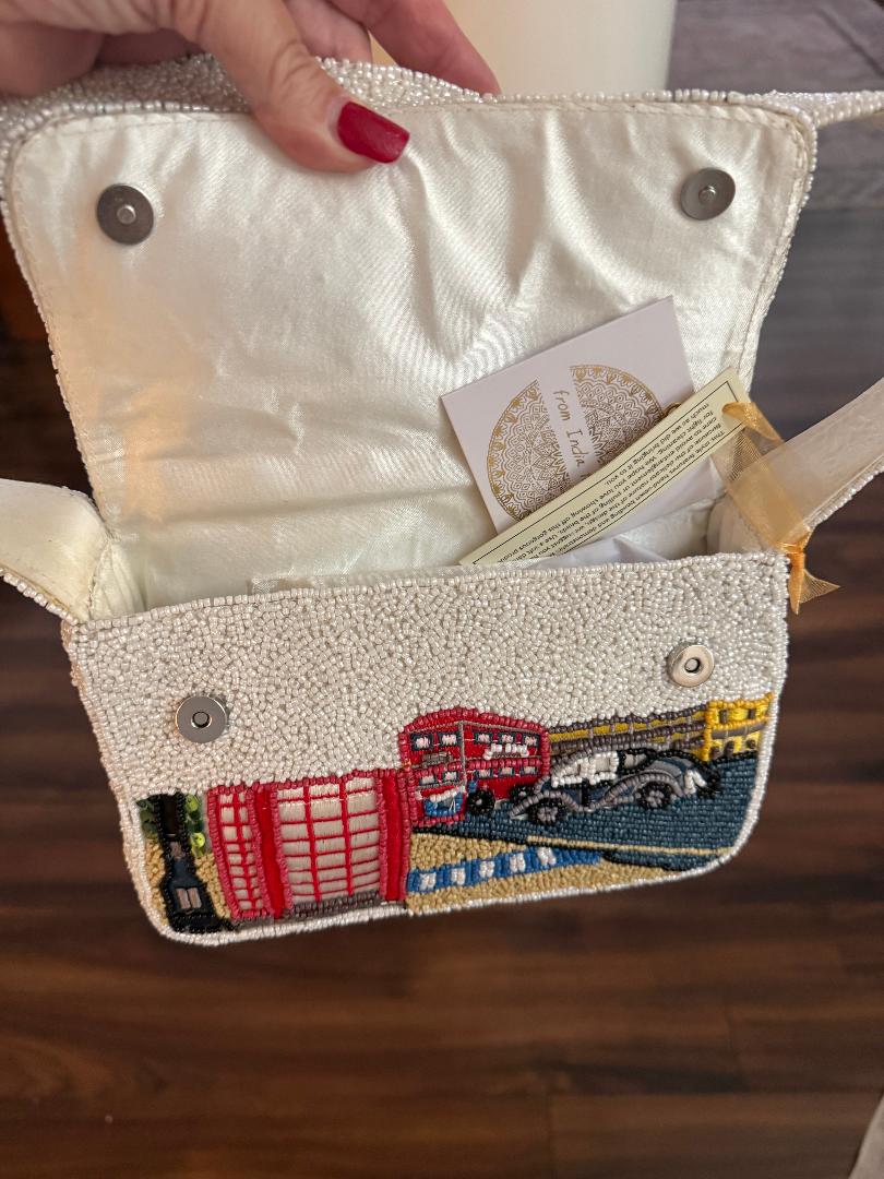 A close-up of a hand holding an open cream-colored beaded clutch bag with embroidered London-themed motifs, including red double-decker buses, a black car, and a red telephone booth, on a wooden floor background.