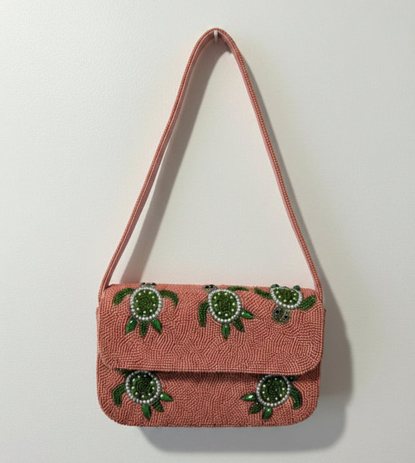 A small pink textured shoulder bag with a green turtle pattern on the front flap, hanging against a plain white wall.