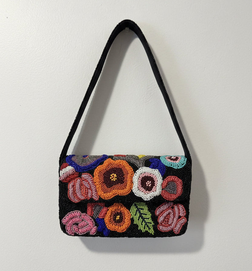 A black shoulder bag with a black strap, decorated with colorful hand-beaded flowers in pink, orange, purple, white, and green on the front flap, shown against a plain light gray background.