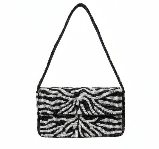 A black beaded and white zebra-print shoulder bag with a black strap, set against a background of reddish-purple and green foliage.