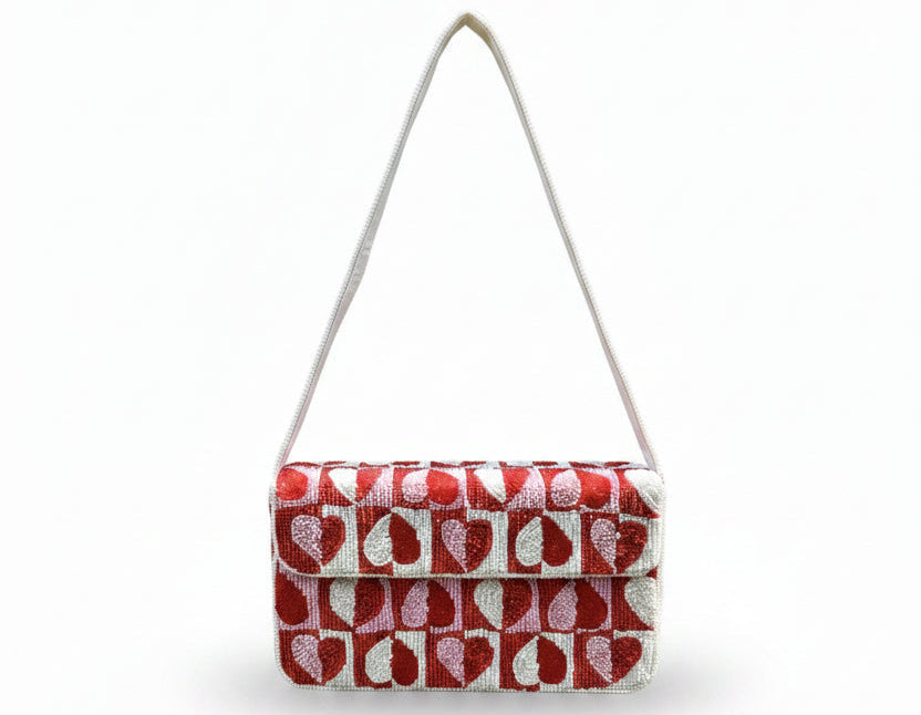 A beaded rectangular shoulder bag with a red and white repeating leaf pattern and a light pink strap, held by a hand against a background of green ferns and trees.