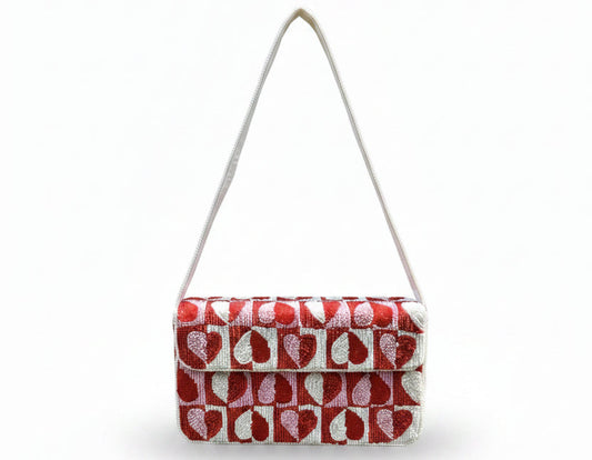 A beaded rectangular shoulder bag with a red and white repeating leaf pattern and a light pink strap, held by a hand against a background of green ferns and trees.