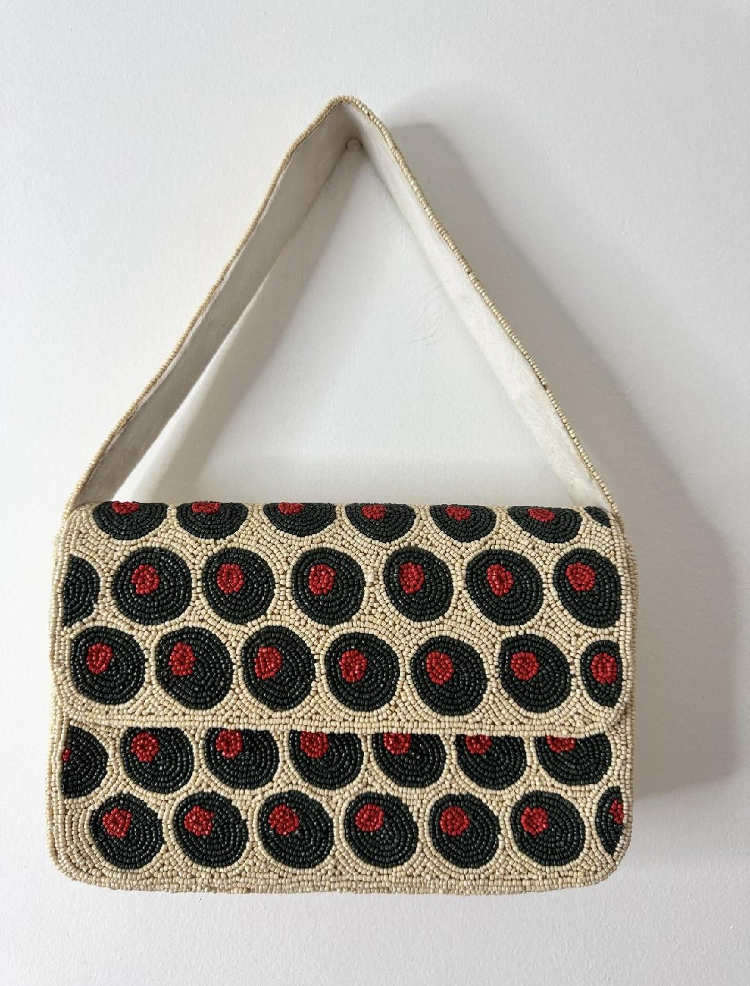 A beaded shoulder bag with a beige base, decorated with rows of black circular motifs containing green olive details, and a light beige strap, shown against a plain white background.