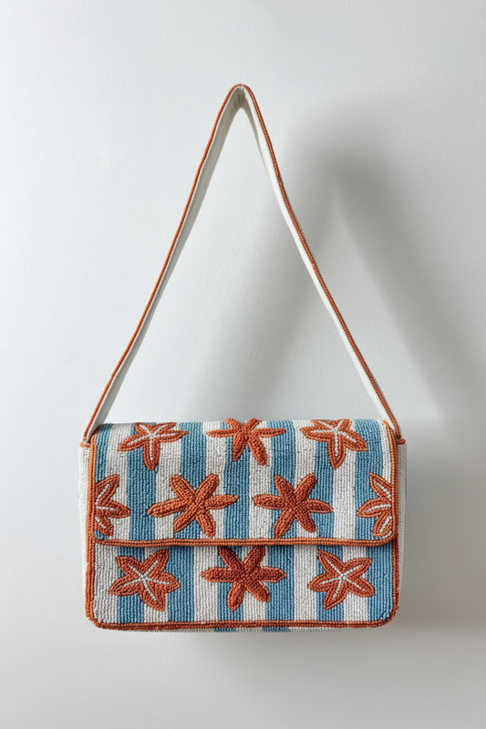A beaded shoulder bag with a blue and white striped body, decorated with orange starburst patterns, and featuring a wide white strap with blue and orange stripes.